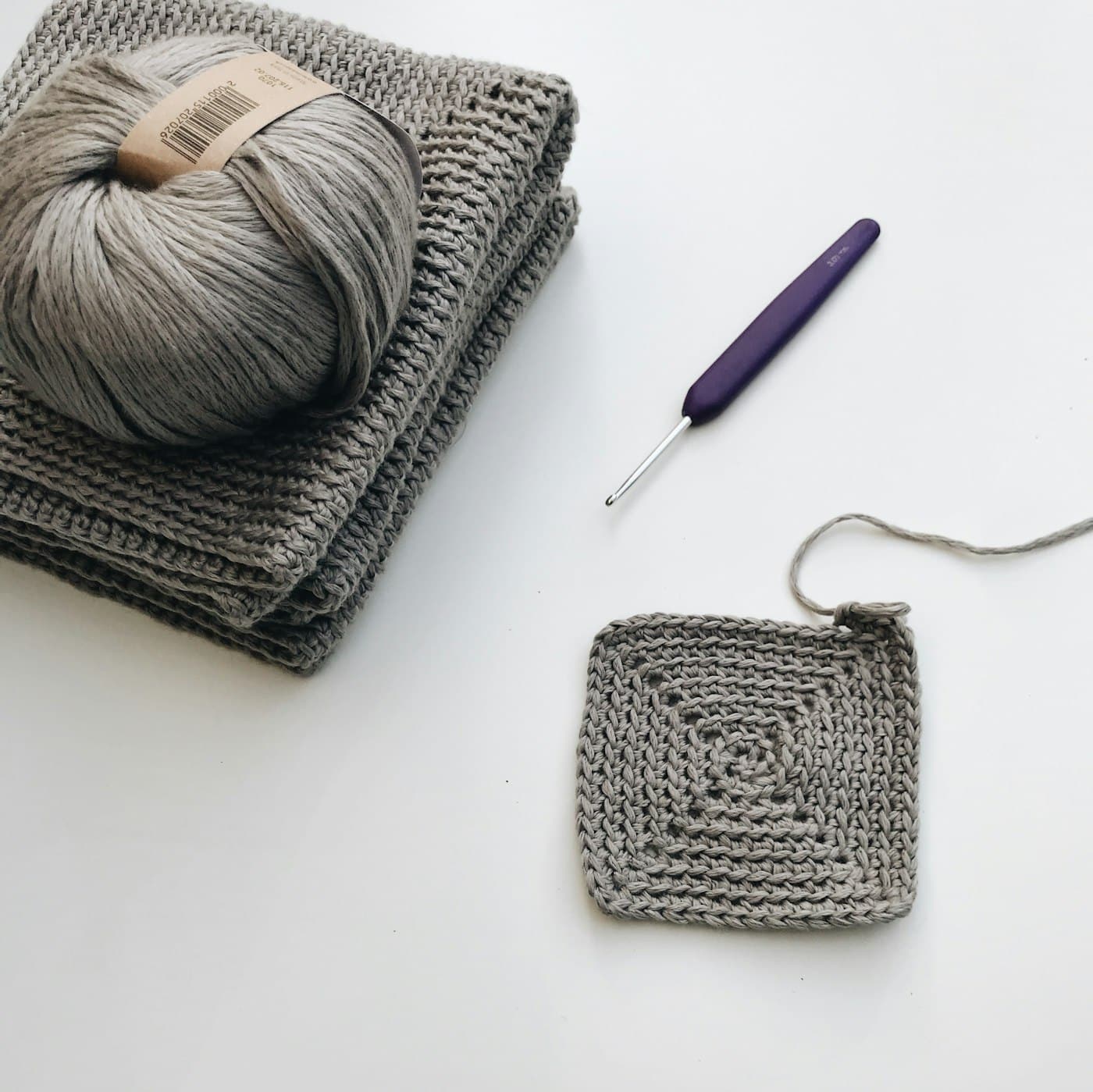 A ball of grey yarn, a crochet hook, and a granny square swatch on a white surface